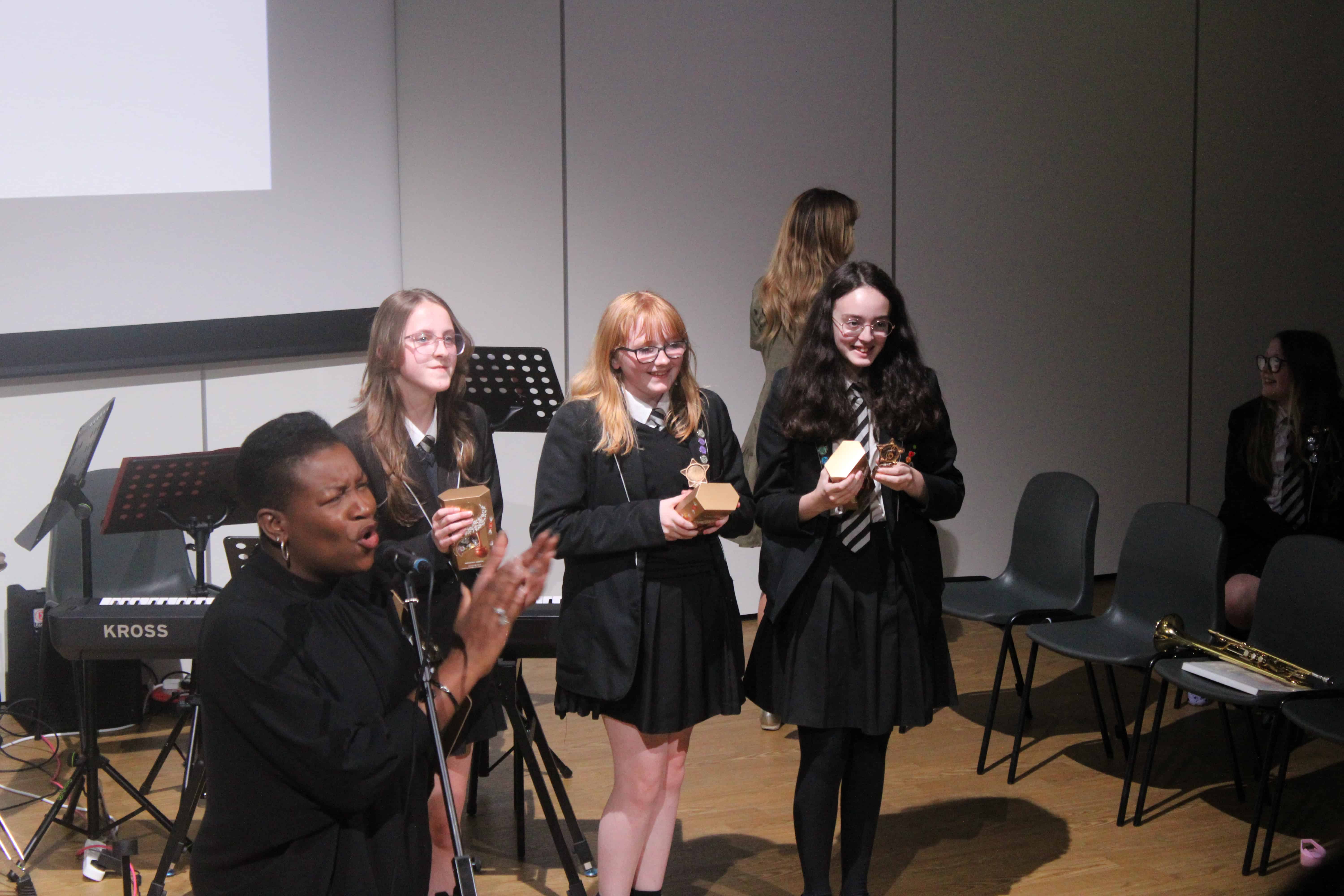 The 3 winning students stand smiling and holding their trophies in front of the Laurus Ryecroft Summer Sounds audience.