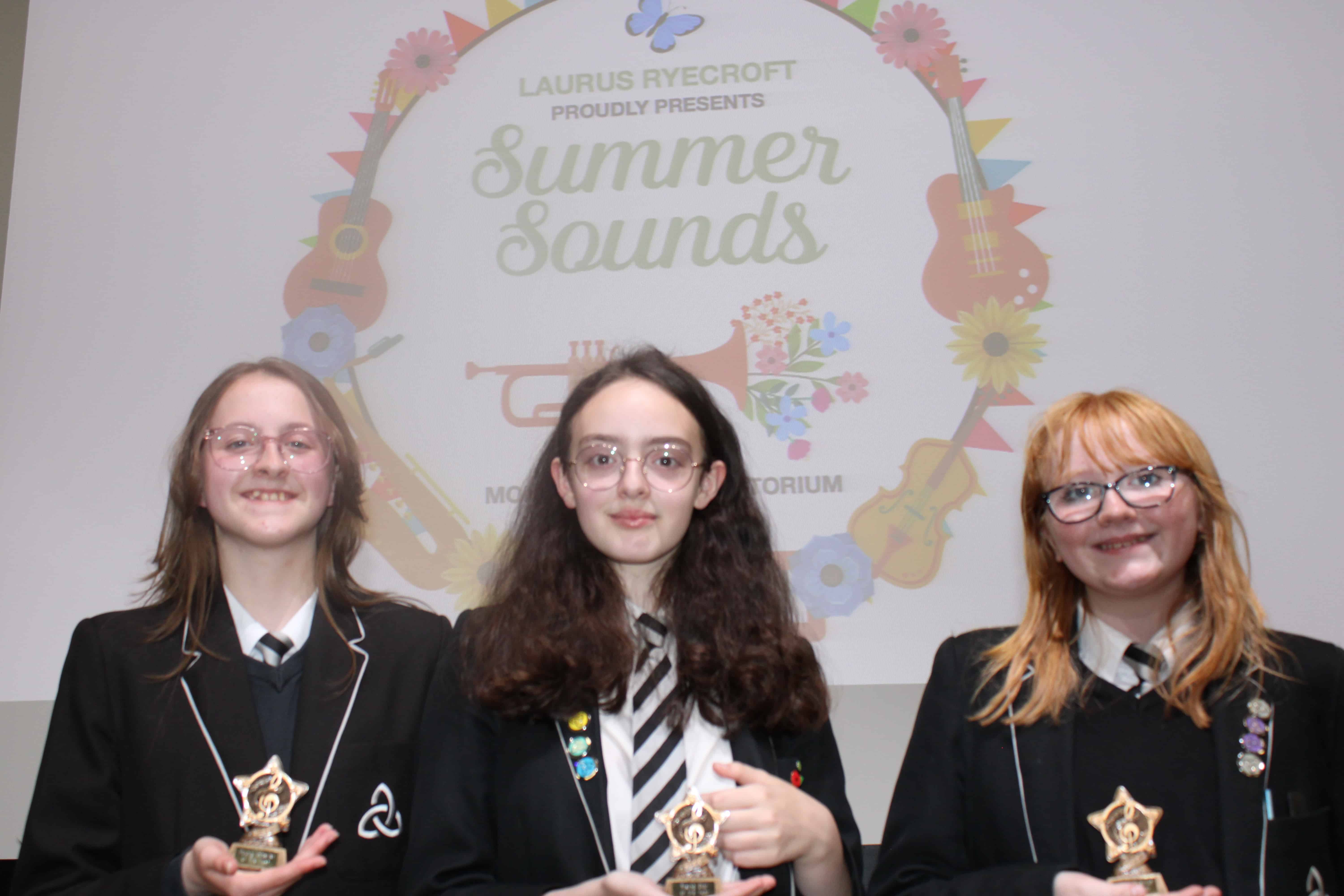 The 3 winning students stand smiling and holding their trophies in front of the Laurus Ryecroft Summer Sounds decorative screen.