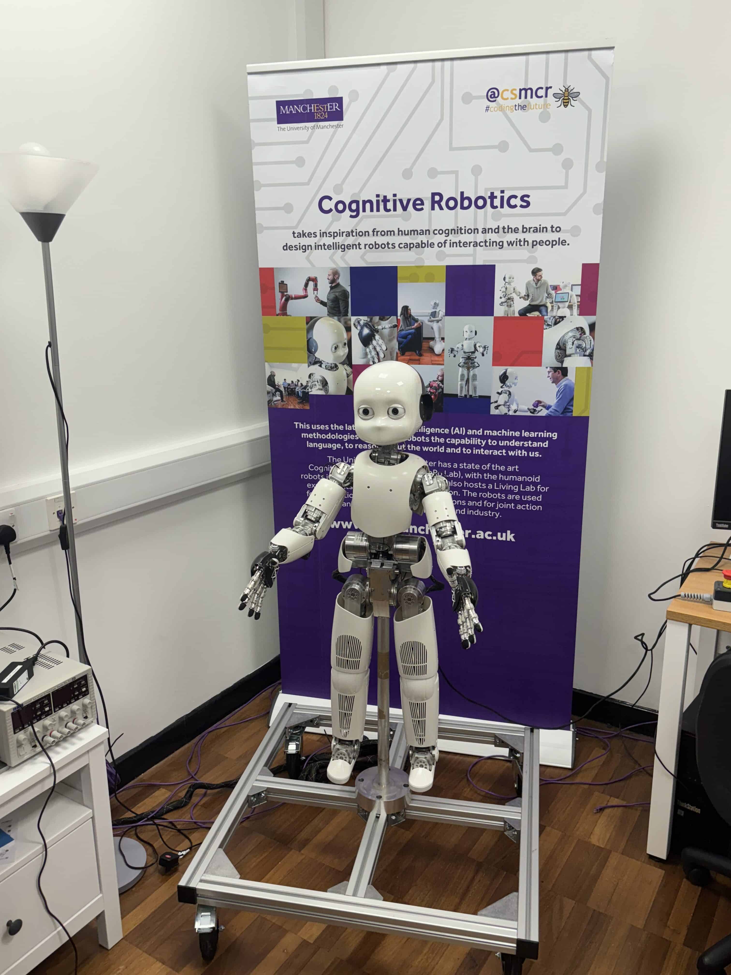A robot in front of a banner about Cognitive Robotics at the University of Manchester during a trip for Laurus Ryecroft students.